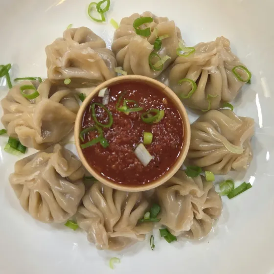 A plate of pleated veg momo around a bowl of house chutney, topped with sliced scallion