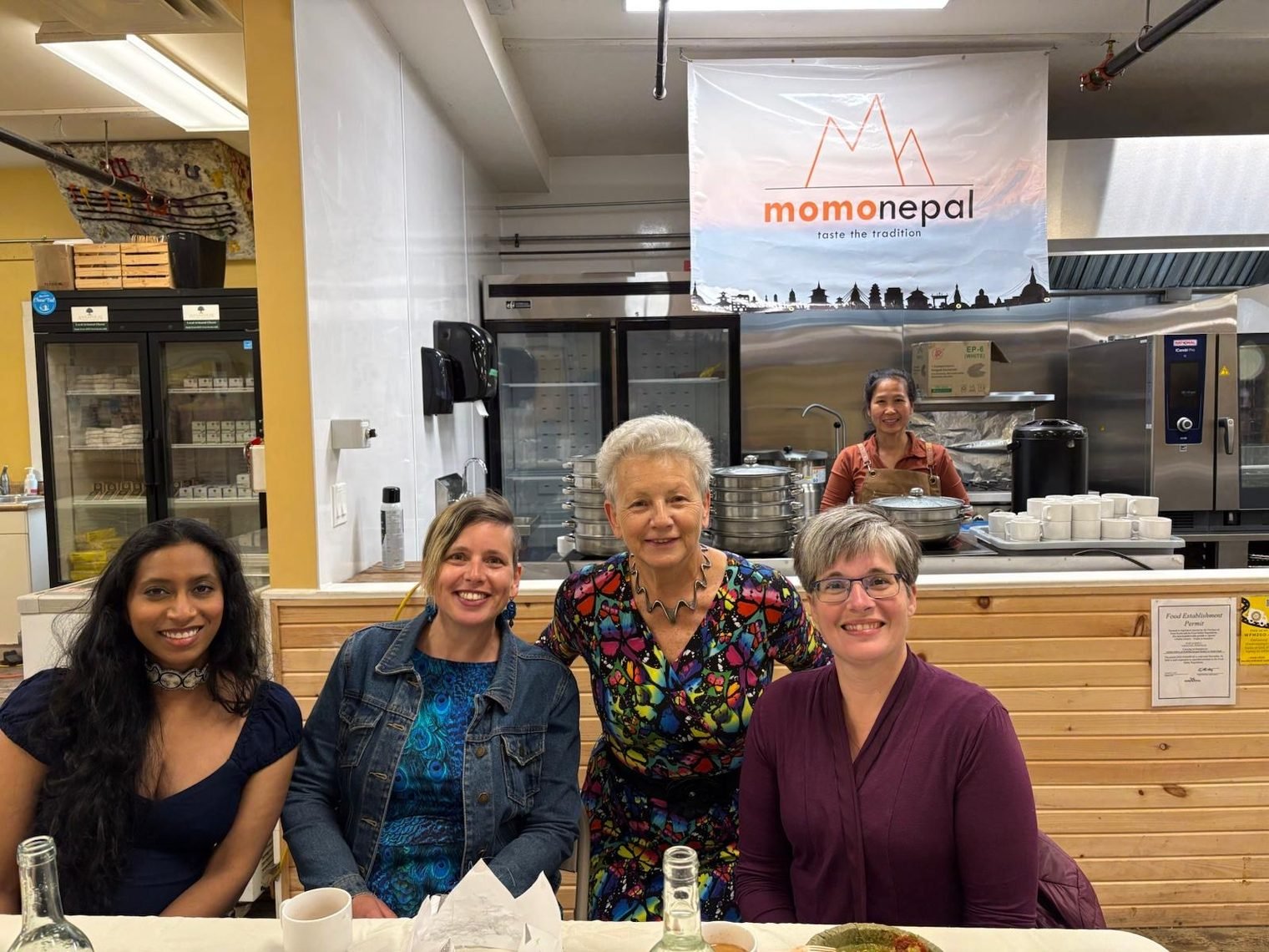 Four guests smiling at a table in front of the MomoNepal counter and banner, with a cook at the counter behind them